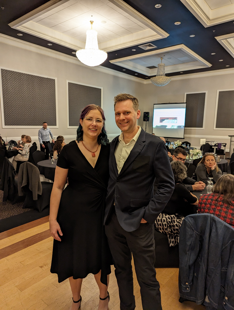 This post features guest speaker Becky Verdun and her partner posing and smiling. Beck wears a knee-length black dress, and her partner wears a grey suit with a white dress shirt.